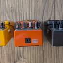 Tři efektové pedály v žluté, oranžové a černé barvě s kovovými ovládacími knoflíky. Každý pedál má na přední straně bílý štítek s nápisem „use BOSS P…