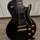 Černá elektrická kytara s gold hardwarem a krémovým lemováním. Má dva humbuckery, čtyři zlaté ovládací knofíky a jeden přepínač. Nástroj je umístěn n…