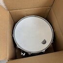 V kartonové krabici se nachází buben s bílým membránou a chromovaným obručem. Na pravé straně membrány je černý oválný štítek s nápisem SONOR, pod ní…