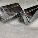 Dva chromované humbucker snímače pro elektrickou kytaru s kovovými kryty. Povrch je lesklý, s viditelnými otisky prstů a drobnými nečistotami. Na sní…
