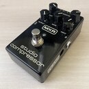 Černý efekt pedál s nápisy studio compressor a logem MXR. Na horní straně jsou čtyři ovládací knoflíky s popisky OUTPUT, RATIO, RELEASE a REDUCTION,…