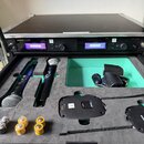 Otevřený černý transportní case s polstřováním, v němž jsou umístěny dva bezdrátové mikrofony a dva vysílače. V horní části je rackový přístroj s náp…