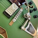 Detail elektrické kytary v matně zelené barvě s hnědým kytarovým popruhem. Na těle je vidět chromovaný Bigsby vibrato mechanismus s nápisem Bigsby®,…
