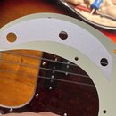 Fotografie zachycuje světlý, krémový pickguard s přilepeným šedým papírovým šablonem. V pickguardu a v šabloně jsou vyvrtané otvory pro potenciometry…