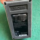 Záběr na zadní stranu efekt pedálu značky BOSS. Na černém plastovém krytu je vidět bílý štítek s nápisy BOSS AC ADAPTOR, specifikacemi napětí a texte…