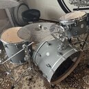 Šedá bicí souprava s kovovým kováním, umístěná na vzorovaném koberci. Na tomu a ostatních bubnech je vidět nápis Epiphone. Souprava obsahuje basový b…