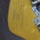 Detail zadní strany hlavy kytary z vybarveného světlého dřeva. Na povrchu je vidět nápis „Made in Korea“ a podpis „Yoshiaki“. V horní části jsou vidě…