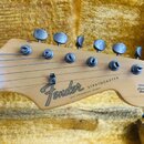 Detail hlavy elektrické kytary z jasného dřeva s nápisy Fender a STRATOCASTER. Pod nimi je menší text WITH STACKSONIZED TECHNOID a vpravo nápis ORIGI…