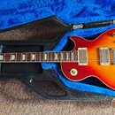Elektrická kytara s trupem v červeno-žlutém přechodovém odstínu (sunburst) umístěná v černém tvrdém kufru s modrým polstrováním. Na hlavě hmatníku je…