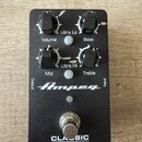 Černý efekt pedál s nápisy Ampeg a CLASSIC Analog Bass Preamp. Zařízení má čtyři černé otočné knoflíky s popisky Volume, Bass, Mid a Treble, dva šedé…