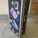 Černý transportní flightcase s hliníkovými hranami a kovovými rohy. Na čelní straně jsou nalepené různé barevné samolepky a nápisy, včetně textu HIER…