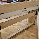 Fotografie zachycuje zadní část digitálního pianina v dřevěném provedení s bílým panelem. Na panelu je umístěno logo a nápis Roland. V dolní části js…