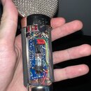 Fotografie zachycuje vnitřek rozebraného mikrofonu s kovovou síťkou a stříbrným povrchem. Uvnitř je vidět modrá tiskovaná spoje s elektronickými souč…