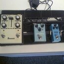 Na černém pedalboardu jsou umístěny tři efektové pedály. Zleva je bílý pedál s nápisem Ibanez, který má čtyři otočné knoflíky a posuvník Delay Time.…