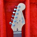 Hlava elektrické kytary z přírodního světlého dřeva v červeném plišovém pouzdře. Na hlavě je černé logo Fender® a nápis STRATOCASTER MADE IN USA. Dol…