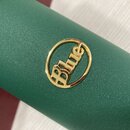 Detailní záběr na zelený povrch s texturou, pravděpodobně pouzdro hudebního nástroje. Uprostřed je umístěn kovový zlatý emblem v kruhu s nápisem Blue…