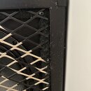 Detailní záběr na roh černého reproduktorového boxu s kovovou mřížkou v kosoctihlavém vzoru. Povrch černého rámu vykazuje známky opotřebení, prach a…
