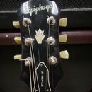 Černý hlavník elektrické kytary s bílými ladicími kolíky. V horní části je nápis Epiphone a uprostřed bílá inkrustace ve tvaru květu. Dole je vidět n…