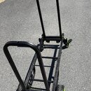 Černý transportní vozík z kovových trubek s čtyřmi koly. Kola mají zelené ráfky a výrazný vzorkovaný plášť. Na rámu jsou vidět zelené detaily v míste…