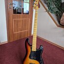 Elektrická basová kytara s korpusem v barevnosti sunburst a černým pickguardem. Na hmatníku jsou vidět zlaté ozdobné vložky a na hlavě nástroje je ná…