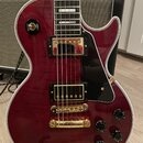 Tělo elektrické kytary v tmavě červené barvě s viditnou strukturou dřeva a bílým lemováním. Nástroj má zlatý hardware, dva humbuckery, čtyři černé ov…