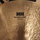 Detailní pohled na povrch mosazného činely s výrazným kovánívým rýhováním. Na povrchu je černý nápis HH Raw Bell Dry Ride 21"/53 cm a nad ním logo SA…