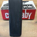Černý kytarový wah pedál s žebrovaným povrchem šlapadla. Na bocích šlapadla jsou vyryty nápisy AMPLIFIER a INSTRUMENT. V pozadí je vidět červeno-bílá…