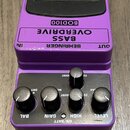 Fialový efekt pedál s černým stoupacím spínačem a ovládacími knoflíky. Na horní části je nápis BEHRINGER, BASS OVERDRIVE a model BOD100. Na spodní čá…