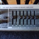 Detail ovládacího panelu šedého zesilovače s nápisem 180-WATT PA SYSTEM/KEYBOARD AMPLIFIER. Panel obsahuje dva otočné knoflíky s popisky FX a LEVEL,…