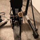 Černý bicí pedál s kovovou konstrukcí a šlapadlem. Na spodní části šlapadla je viditelné logo Pearl. K pedálu je připojena kulatá kartáčová koštěčka.…