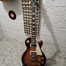 Elektrická kytara s trupem v tmavě červeném sunburst provedení a bílým pickguardem. Na hlavě hmatníku je nápis Epiphone, pod ním model Les Paul a dál…