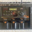 Černý kytarový syntetizátor Roland GR-33 Guitar Synthesizer. Zařízení má digitální displej s nápisem Roland GR-33 Guitar Synth, několik ovládacích tl…