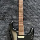 Elektrická kytara s černým bodies, hnědým hmatníkem a světle hnědou hlavou. Na hlavě je nápis Fender Stratocaster. Kytara má tři bílé snímače, tři bí…