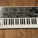 Bílý syntetizátor s klávesnicí a ovládacím panelem v horní části. Na levé straně je nápis Roland GAIA SYNTHESIZER SH-01. Ovládací panel obsahuje různ…