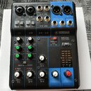 Kompaktní míchací pult Yamaha MG06 v černé barvě s modrými prvky. Na horní části jsou vstupy a výstupy s nápisy 1/L MIC / LINE, 2/R MIC / LINE, L/MON…