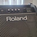 Černý osobní monitor s kovovou mřížkou, na které je bílý nápis Roland. Horní ovládací panel obsahuje vstupy INPUT (V-Drums, LINE), ovladače VOLUME (V…