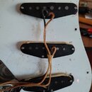 Detailní pohled na bílý pickguard elektrické kytary se třemi černými snímači. Povrch plastu je znečištěný a obsahuje hnědé skvrny. Mezi snímači jsou…