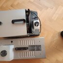 Dvě stříbrné kovové krabice s černými madly, položené na dřevěné podlaze. Horní zařízení má otevřenou boční část, kde je vidět bílá plastová nádoba a…