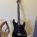 Černá elektrická kytara s tmavým hmatníkem a bílým pickguardem. Na hlavě je bílý nápis "Fender" a menší text "STRATOCASTER". Nástroj vykazuje známky…