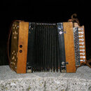Dřevěný harmonika s černým měchem a hnědými koženými popruhy. Na pravé straně jsou vidět bílé tlačítka a kovová mřížka. Povrch dřeva vykazuje známky…