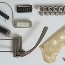 Sada různých kovových a plastových komponentů pro kytaru na bílém pozadí. V horní části je černý snímač s nápisem BRILANT DE LUXE, kovová hlava s lad…