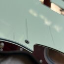 Detailní záběr na světle zelený plastový pickguard kytary. Povrch vykazuje několik viditelných škrábanců a oděrů. V dolní části je vidět otvor pro šr…