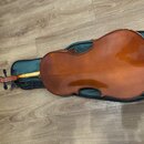 Hnědé cello s lakovaným povrchem položené na tmavě zeleném obalu. Nástroj má tradiční tvar s viditelným hmatníkem a ladicími kolíky. Na fotce nejsou…