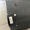 Detailní záběr na černý kout s texturovaným povrchem, pravděpodobně součást komba nebo reproduktoru. Na černém plastovém rohu je nalepený bílý štítek…