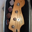 Hlava basové kytary z přírodního dřeva v černém tvrdém kufru. Na dřevě jsou nápisy Fender, JAZZ BASS® a MADE IN MEXICO. V horní části je svislý nápis…
