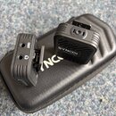 Na černém pouzdře leží dva černé bezdrátové vysílače značky SYNCO. Pravý jednotka nese nápis SYNCO Transmitter, levá má označení OUT a symbol krucové…