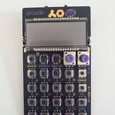 Černý elektronický sekvencer s displejem a ovládacími prvky. V horní části je nápis arcade, logo XO a nálepka CHEAP MONDAY. Zařízení obsahuje řadu tl…