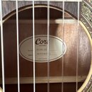 Detail pohledu do ozvučnice akustické kytary. Uvnitř je nalepený bílý oválný štítek s nápisy Cort GUITARS, MODEL AC100 SG, SERIAL NO IA2503819 01 a M…