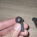 Ruka drží jedno bezdrátové sluchátko v tmavě šedé barvě. Na spodní části tětěnky jsou vidět dva kovové kontakty pro nabíjení. Sluchátko má nasazenou…