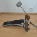 Kovový basový pedál s černou protiskluzovou plochou na stupni. Beater je zakončen šedým plstěným nebo vlněným klubíkem. Na mechanismu je vidět hnědý…
